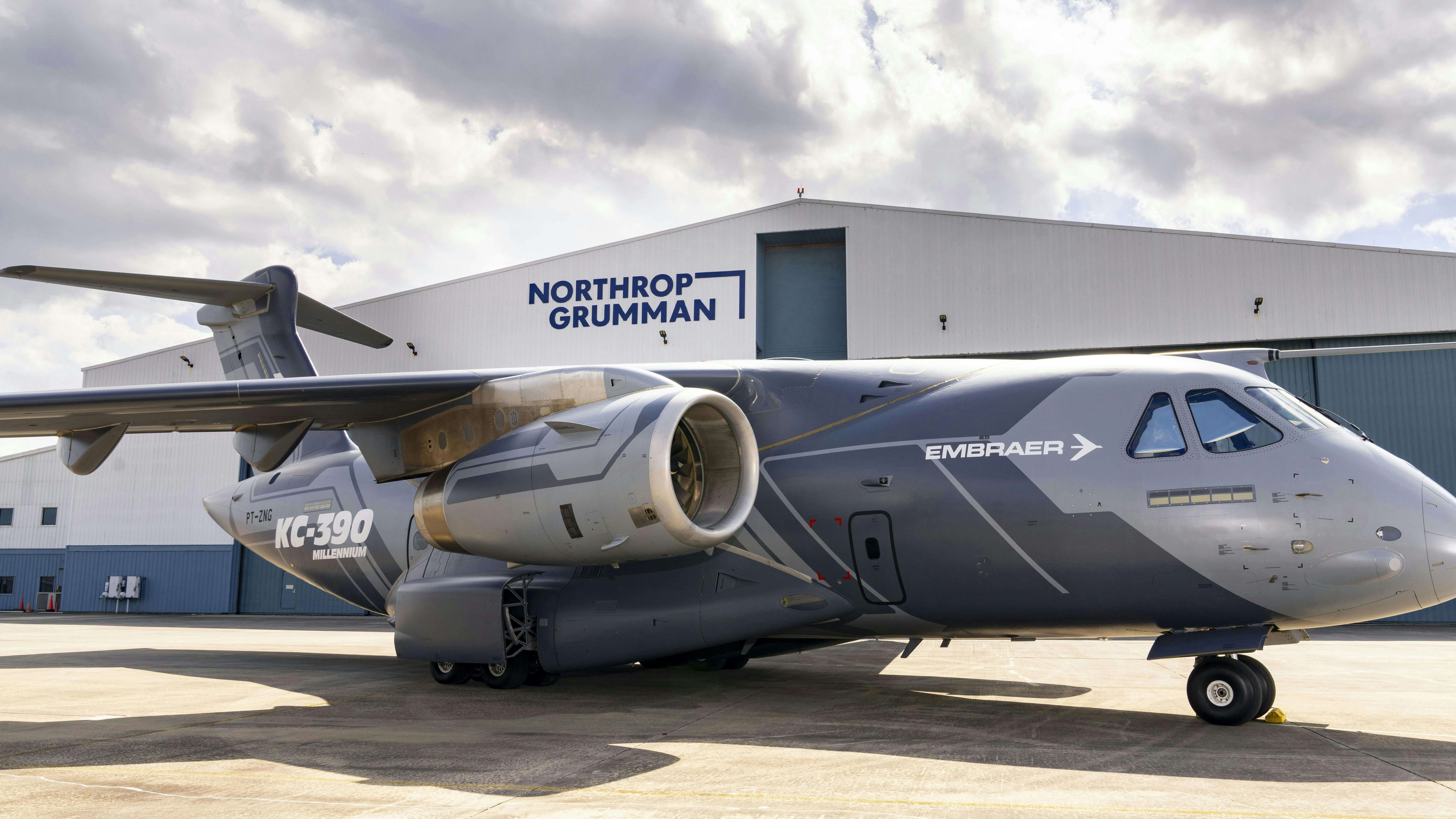 A gray airplane with text on it that reads 'EMRAER' and 'KC-390 Millennium' parked in front of a hangar with a sign on it that reads, 'Northrop Grumman'