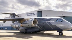 A gray airplane with text on it that reads 'EMRAER' and 'KC-390 Millennium' parked in front of a hangar with a sign on it that reads, 'Northrop Grumman' A gray airplane with text on it that reads 'EMRAER' and 'KC-390 Millennium' parked in front of a hangar with a sign on it that reads, 'Northrop Grumman'
