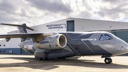A gray airplane with text on it that reads 'EMRAER' and 'KC-390 Millennium' parked in front of a hangar with a sign on it that reads, 'Northrop Grumman' A gray airplane with text on it that reads 'EMRAER' and 'KC-390 Millennium' parked in front of a hangar with a sign on it that reads, 'Northrop Grumman'