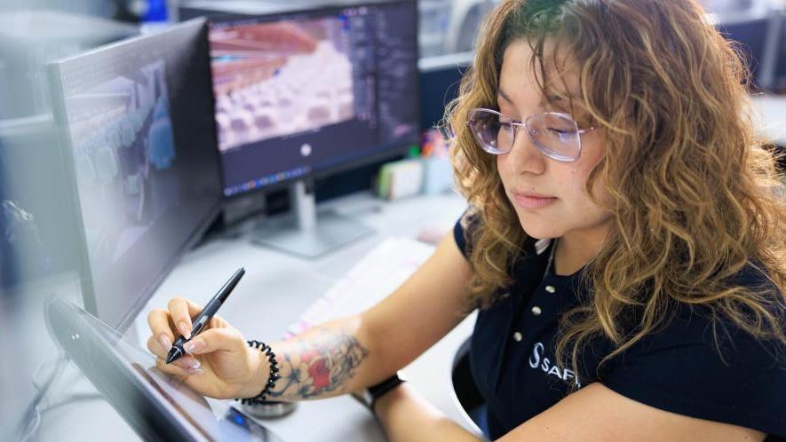 A woman wearing a shirt that says, 'SAFRAN' while sitting at a desk and writing on a tablet with a stylus, with two desktop computers behind her