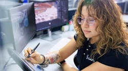 A woman wearing a shirt that says, 'SAFRAN' while sitting at a desk and writing on a tablet with a stylus, with two desktop computers behind her A woman wearing a shirt that says, 'SAFRAN' while sitting at a desk and writing on a tablet with a stylus, with two desktop computers behind her
