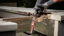 A pair of hands wearing black gloves using a red and white saw blade to cut through a piece of wood A pair of hands wearing black gloves using a red and white saw blade to cut through a piece of wood