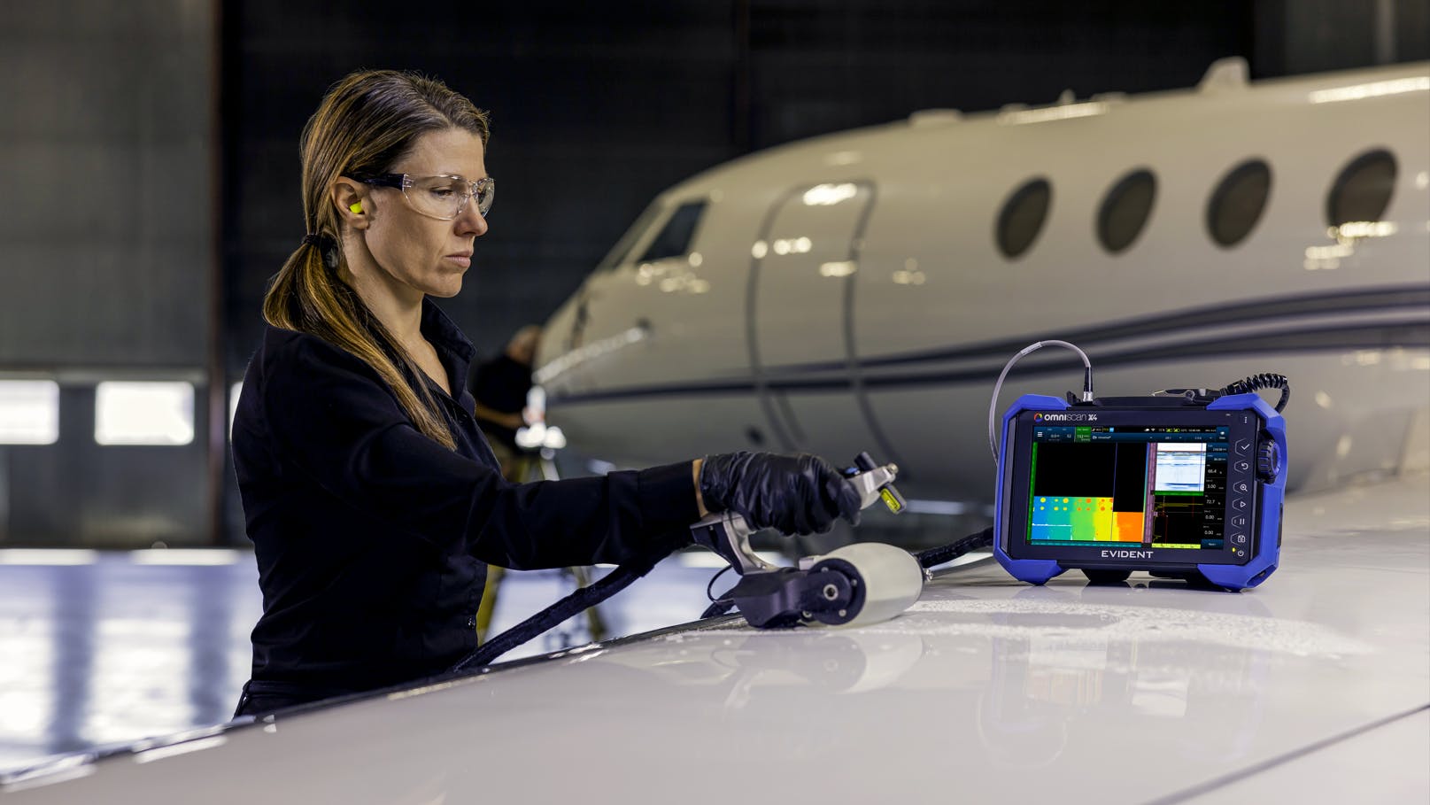 A woman wearing gloves and other PPE using a non-destructive testing device to inspect an airplane, with a handheld part and a screen