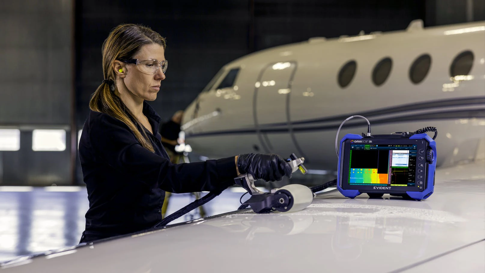 A woman wearing gloves and other PPE using a non-destructive testing device to inspect an airplane, with a handheld part and a screen