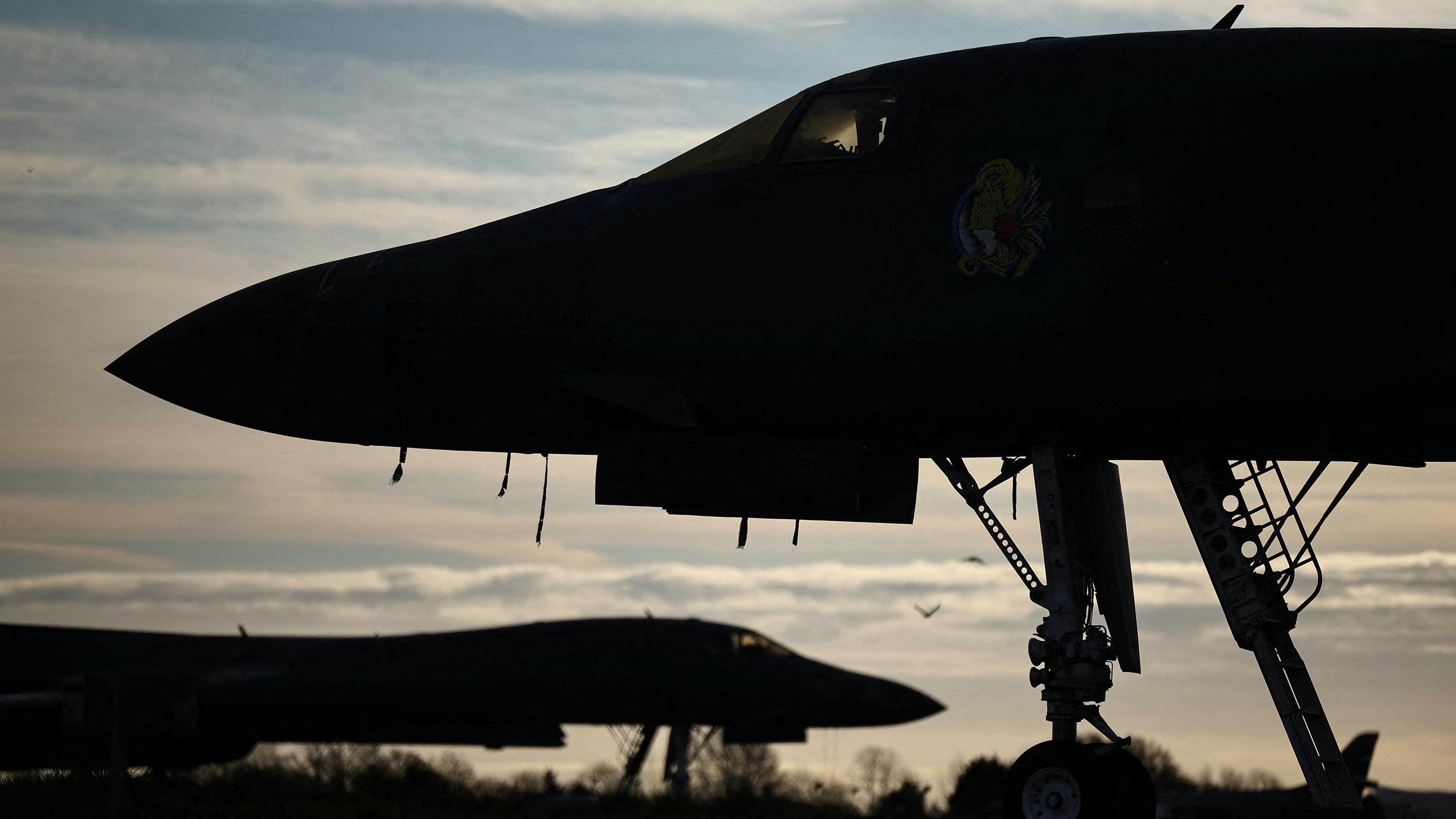 A silhouette of two aircraft standing near trees against a pale blue sky with clouds