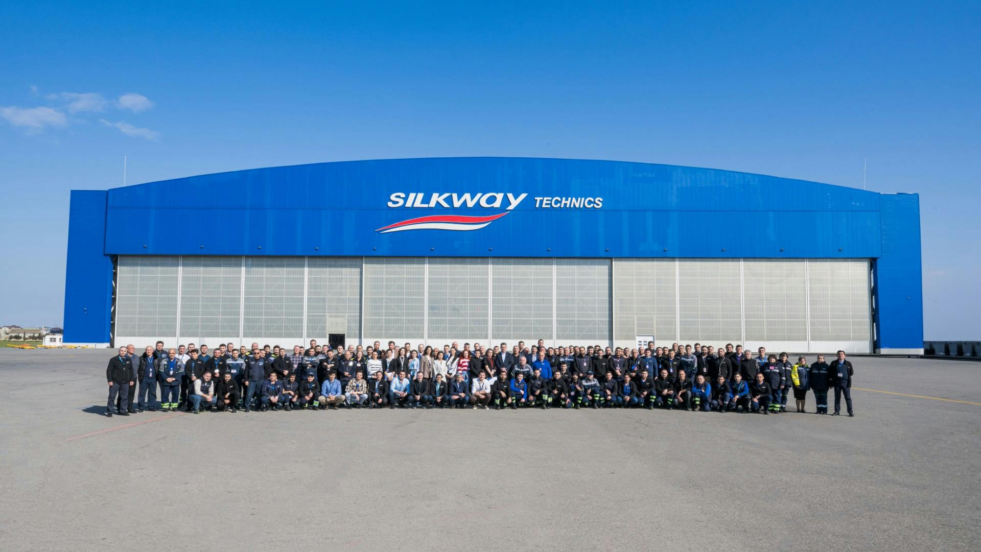 A large crowd of people gathering for a group photo in front of a large blue building with 3 hangar doors and a sign that reads: Silk Way Technics