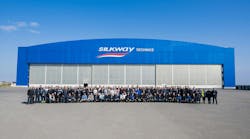 A large crowd of people gathering for a group photo in front of a large blue building with 3 hangar doors and a sign that reads: Silk Way Technics A large crowd of people gathering for a group photo in front of a large blue building with 3 hangar doors and a sign that reads: Silk Way Technics