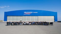 A large crowd of people gathering for a group photo in front of a large blue building with 3 hangar doors and a sign that reads: Silk Way Technics A large crowd of people gathering for a group photo in front of a large blue building with 3 hangar doors and a sign that reads: Silk Way Technics