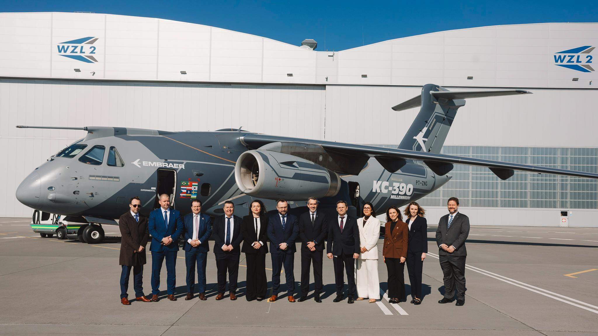 A group of professionals wearing business attire standing in front of a gray aircraft with 'Embraer KC-390 Millennium' printed on the side, with a building in the background labeled 'WZL-2'
