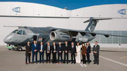 A group of professionals wearing business attire standing in front of a gray aircraft with 'Embraer KC-390 Millennium' printed on the side, with a building in the background labeled 'WZL-2'