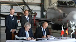 Two men wearing suits standing behind two more men wearing suits sitting down at a table and signing papers, with an airplane parked behind them inside a hangar Two men wearing suits standing behind two more men wearing suits sitting down at a table and signing papers, with an airplane parked behind them inside a hangar