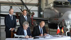Two men wearing suits standing behind two more men wearing suits sitting down at a table and signing papers, with an airplane parked behind them inside a hangar Two men wearing suits standing behind two more men wearing suits sitting down at a table and signing papers, with an airplane parked behind them inside a hangar