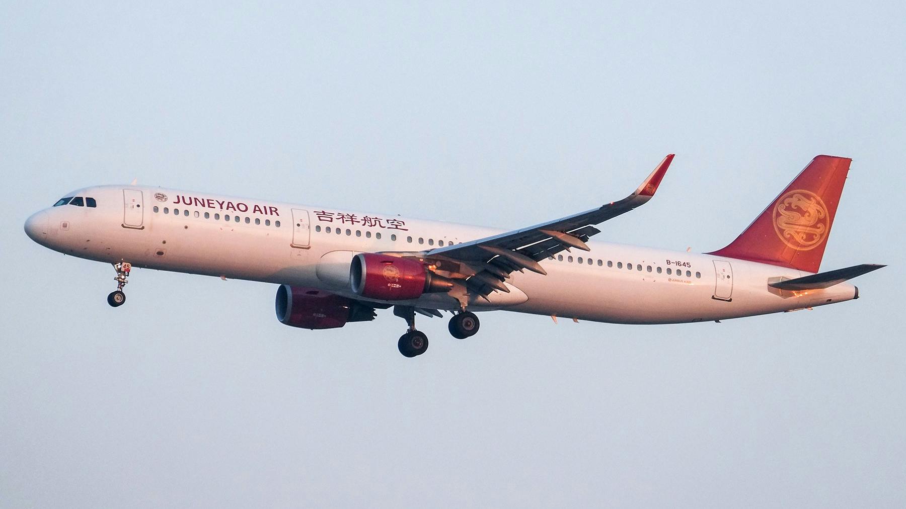 A white airplane with red accents on the engines, wings and tail with text printed on the side in Chinese characters and English text that reads: Juneyao Air