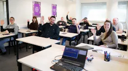 A group of people sitting in a classroom at desks while smiling and waving to the camera