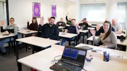 A group of people sitting in a classroom at desks while smiling and waving to the camera