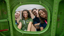 Four people wearing polo shirts and ID badges looking through the window of a green airplane wall smiling at the camera