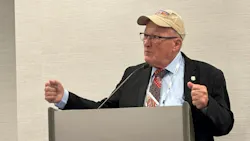 A man wearing business attire and a baseball cap standing at a podium with his arms raised, speaking into a microphone