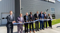 A line of professionals wearing business attire smiling at the camera while standing by a large ribbon during a ribbon-cutting ceremony