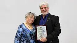 A man and women in professional attire smiling at the camera while standing close to each other, with the man holding a plaque