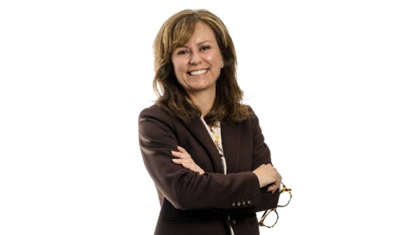 A woman wearing a brown suit and holding a pair of eyeglasses smiling at the camera