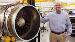 A man wearing khakis and a button-down shirt smiling while leaning on an aircraft engine held in an engine stand