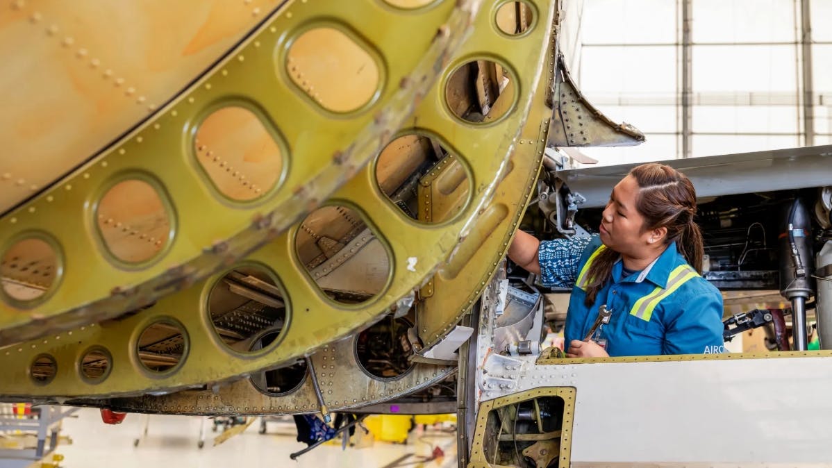 A woman wearing a blue shirt and high-visibility vest working on an airplane that's showing signs of rust