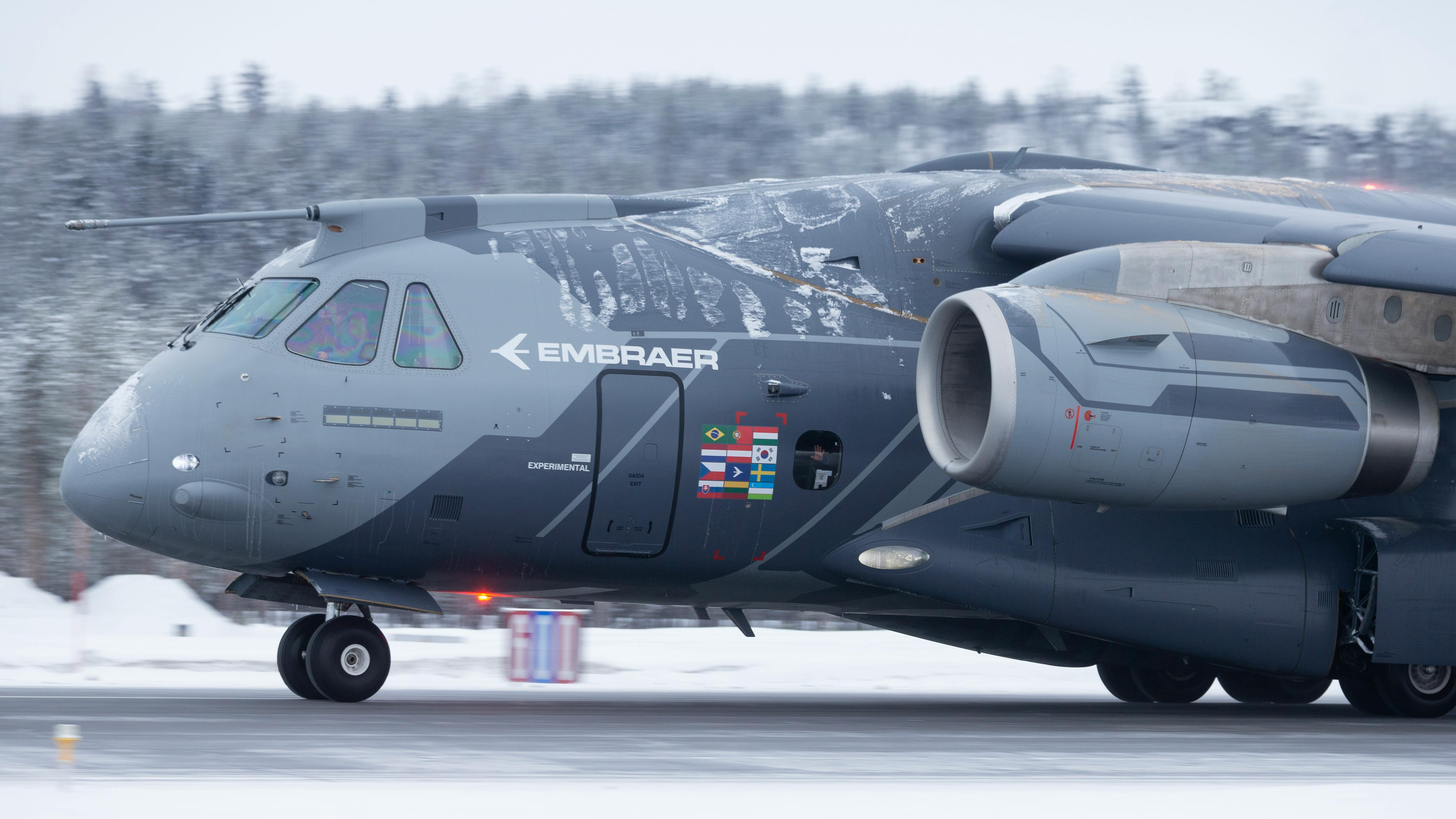 A large aircraft with the word 'EMBRAER' printed on the side parked on the runway with snow on the ground and trees in the background