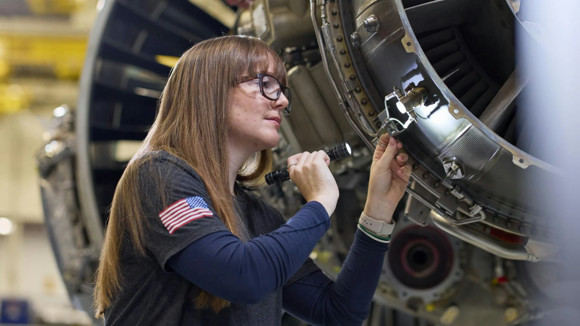 A woman shining a flashlight on an airplane engine and inspecting it carefully