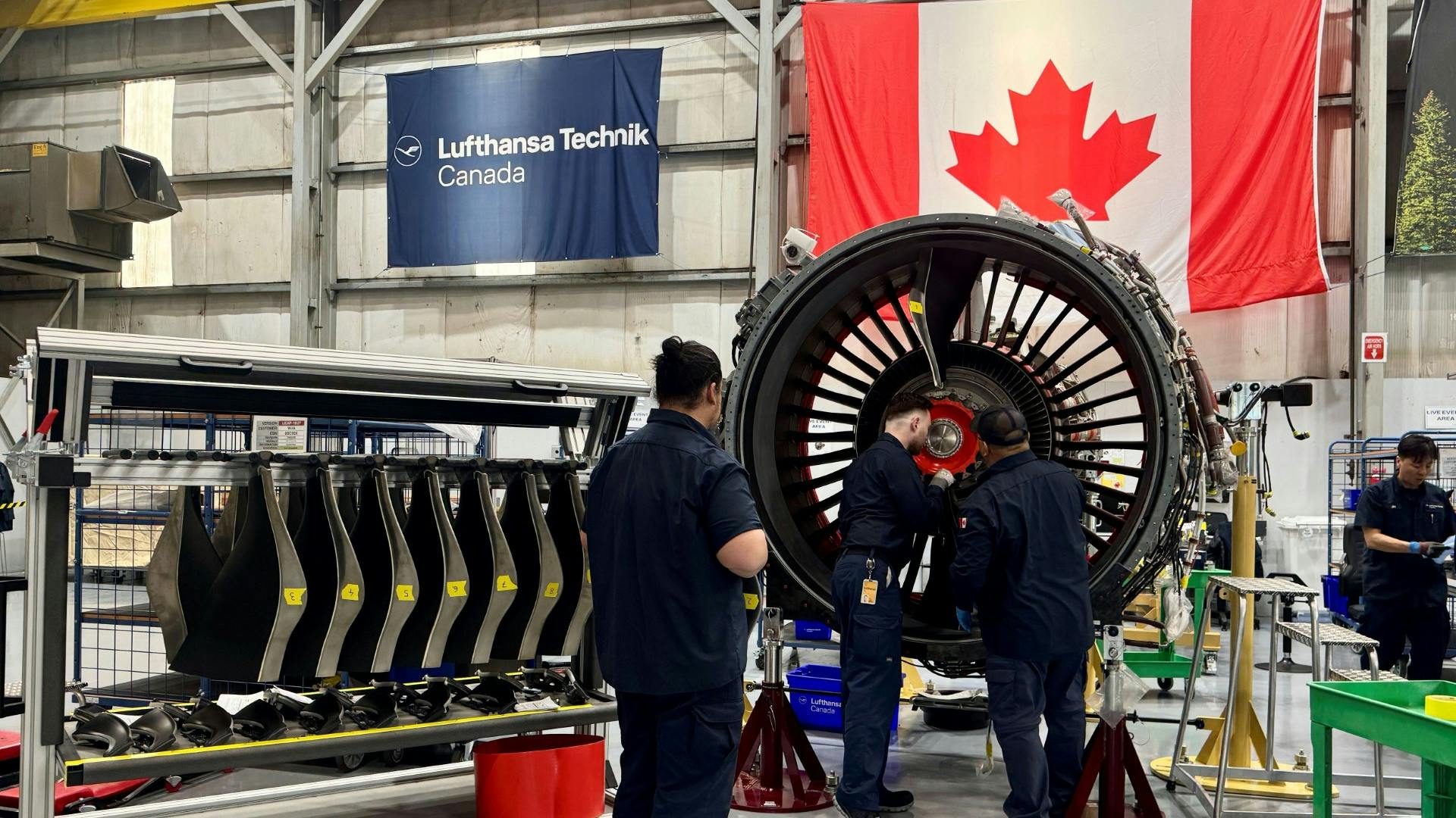 Three aviation maintenance technicians working on a large, black airplane engine, with a Canadian flag and a banner that reads 'Lufthansa Technik Canada' behind the engine