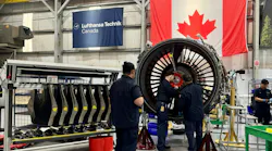 Three aviation maintenance technicians working on a large, black airplane engine, with a Canadian flag and a banner that reads 'Lufthansa Technik Canada' behind the engine Three aviation maintenance technicians working on a large, black airplane engine, with a Canadian flag and a banner that reads 'Lufthansa Technik Canada' behind the engine
