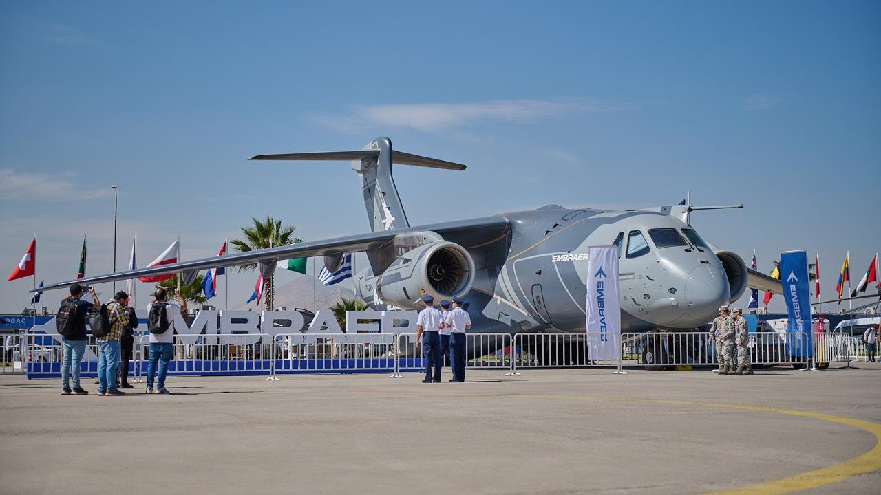 A large military aircraft on display behind a fence, with flags behind the aircraft and a sign that says 'EMBRAER' in front of it