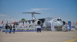A large military aircraft on display behind a fence, with flags behind the aircraft and a sign that says 'EMBRAER' in front of it A large military aircraft on display behind a fence, with flags behind the aircraft and a sign that says 'EMBRAER' in front of it