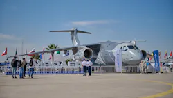 A large military aircraft on display behind a fence, with flags behind the aircraft and a sign that says 'EMBRAER' in front of it A large military aircraft on display behind a fence, with flags behind the aircraft and a sign that says 'EMBRAER' in front of it