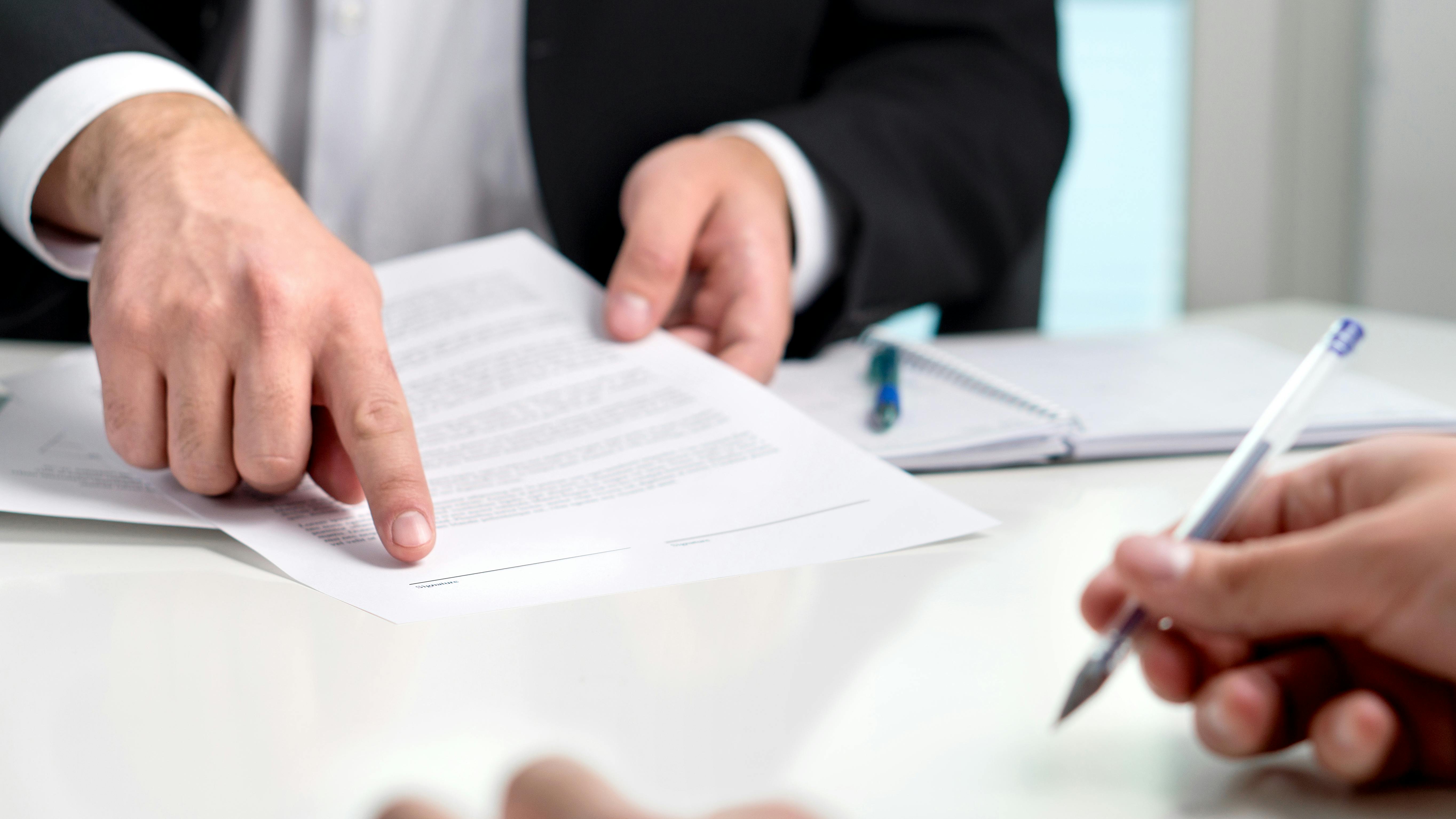 A close-up shot of a table with a person wearing a suit holding a paper and pointing and another person writing