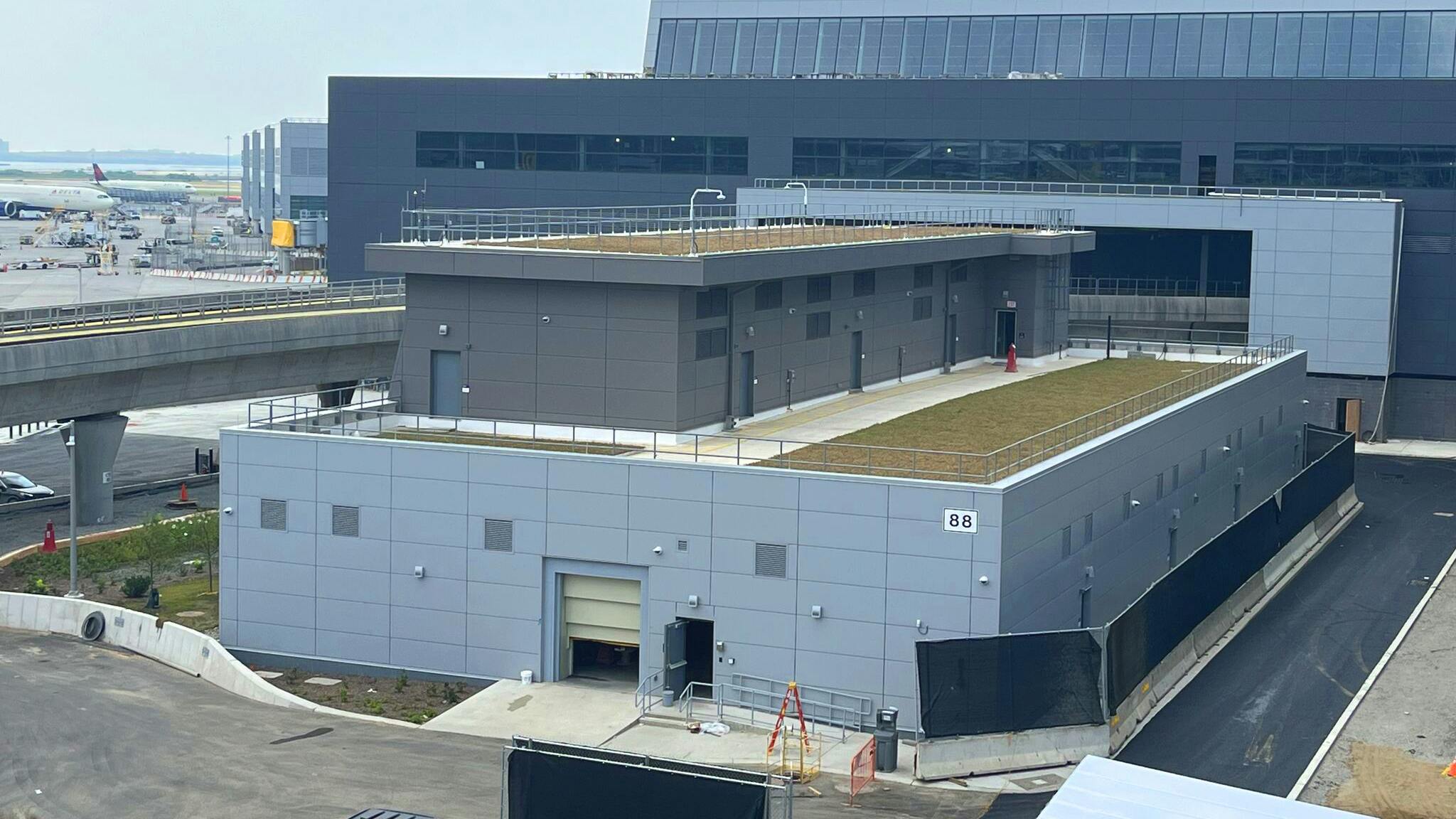 CS2 is in front, New Terminal One is in the background, and AirTrain is on the left. As a 2-story structure with green roof and textured metal panels aligned, the substation blends seamlessly into the airport&rsquo;s modern landscape.