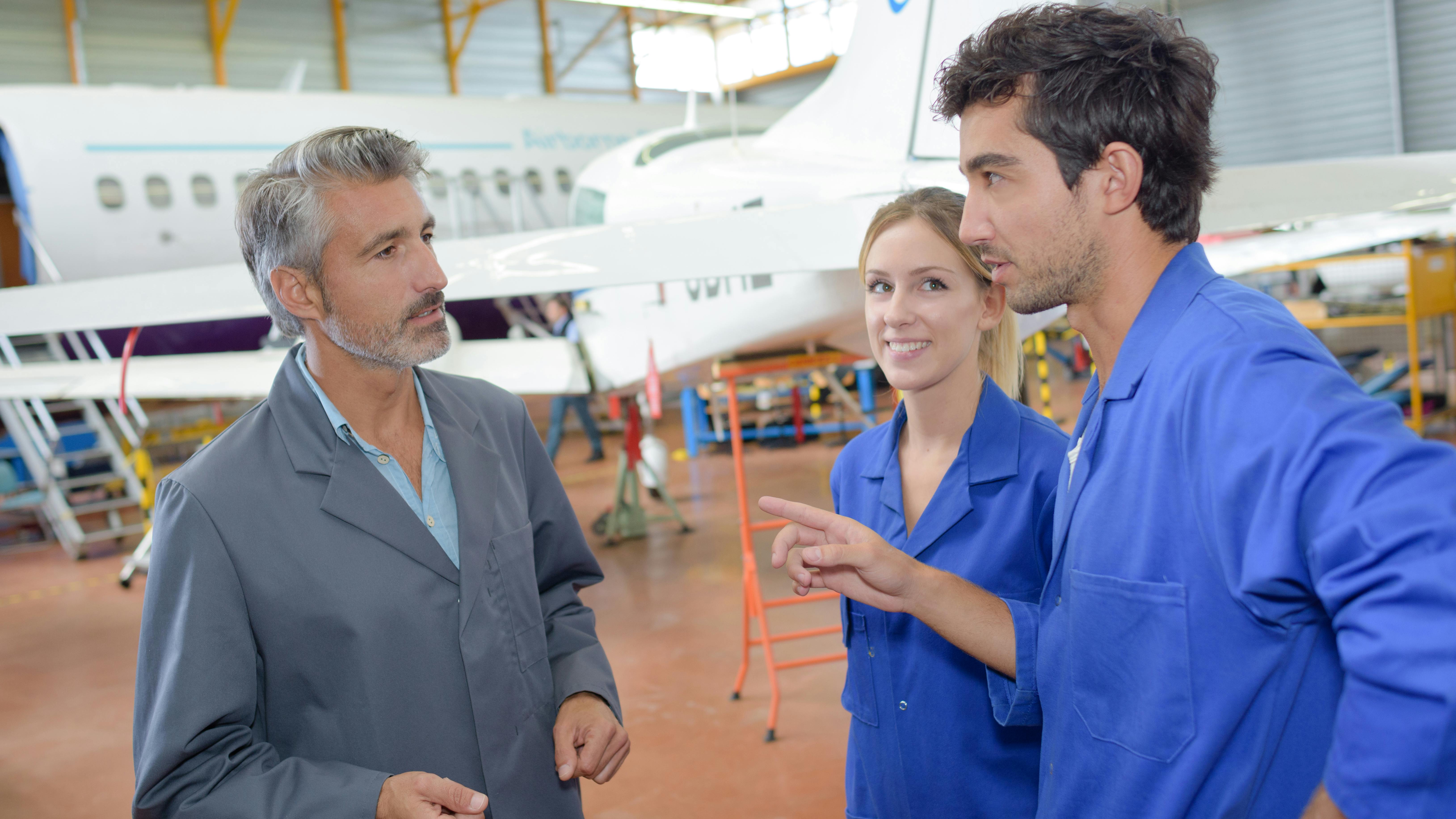 An older aircraft maintenance technician talking to two younger technicians, all three standing in a maintenance hangar in front of a parked airplane