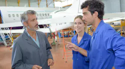 An older aircraft maintenance technician talking to two younger technicians, all three standing in a maintenance hangar in front of a parked airplane An older aircraft maintenance technician talking to two younger technicians, all three standing in a maintenance hangar in front of a parked airplane