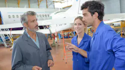 An older aircraft maintenance technician talking to two younger technicians, all three standing in a maintenance hangar in front of a parked airplane An older aircraft maintenance technician talking to two younger technicians, all three standing in a maintenance hangar in front of a parked airplane