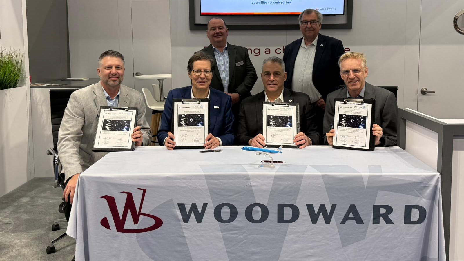 A group of professionals wearing suits holding clipboards and smiling at the camera while gathering around a table sporting a tablecloth with the word 'WOODWARD' on it