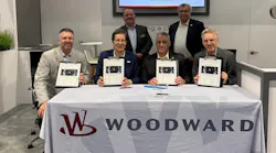 A group of professionals wearing suits holding clipboards and smiling at the camera while gathering around a table sporting a tablecloth with the word 'WOODWARD' on it A group of professionals wearing suits holding clipboards and smiling at the camera while gathering around a table sporting a tablecloth with the word 'WOODWARD' on it