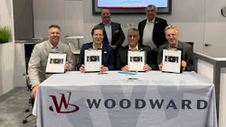 A group of professionals wearing suits holding clipboards and smiling at the camera while gathering around a table sporting a tablecloth with the word 'WOODWARD' on it A group of professionals wearing suits holding clipboards and smiling at the camera while gathering around a table sporting a tablecloth with the word 'WOODWARD' on it
