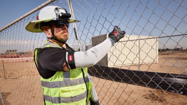 A man wearing a hard hat, high-visibility vest, and protective sleeves standing on a work site near a chain-link fence