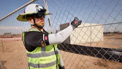A man wearing a hard hat, high-visibility vest, and protective sleeves standing on a work site near a chain-link fence A man wearing a hard hat, high-visibility vest, and protective sleeves standing on a work site near a chain-link fence
