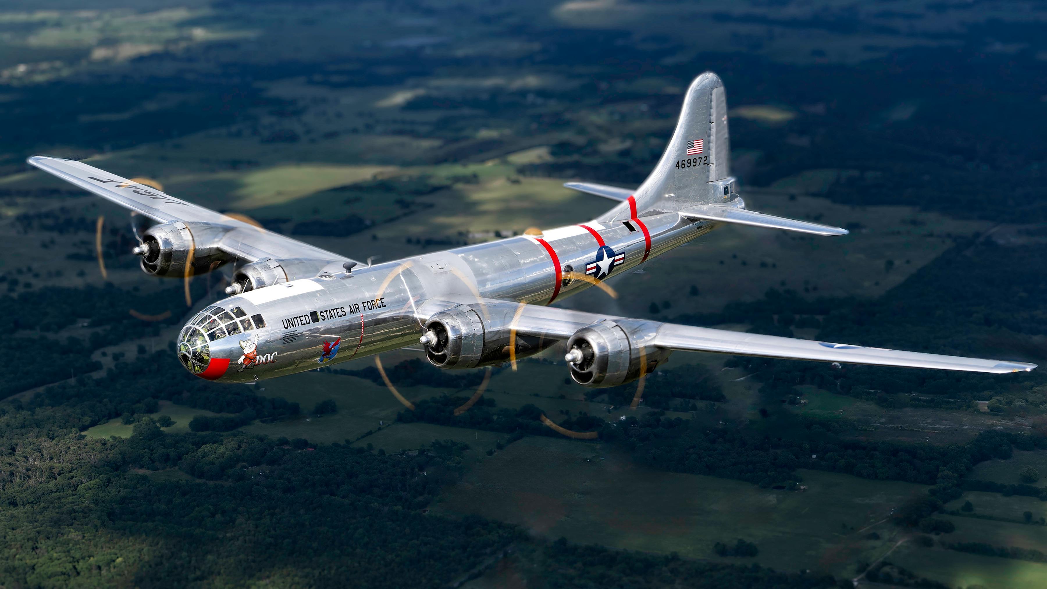 A silver military airplane with red accents flying over lush green land