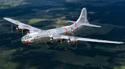 A silver military airplane with red accents flying over lush green land A silver military airplane with red accents flying over lush green land