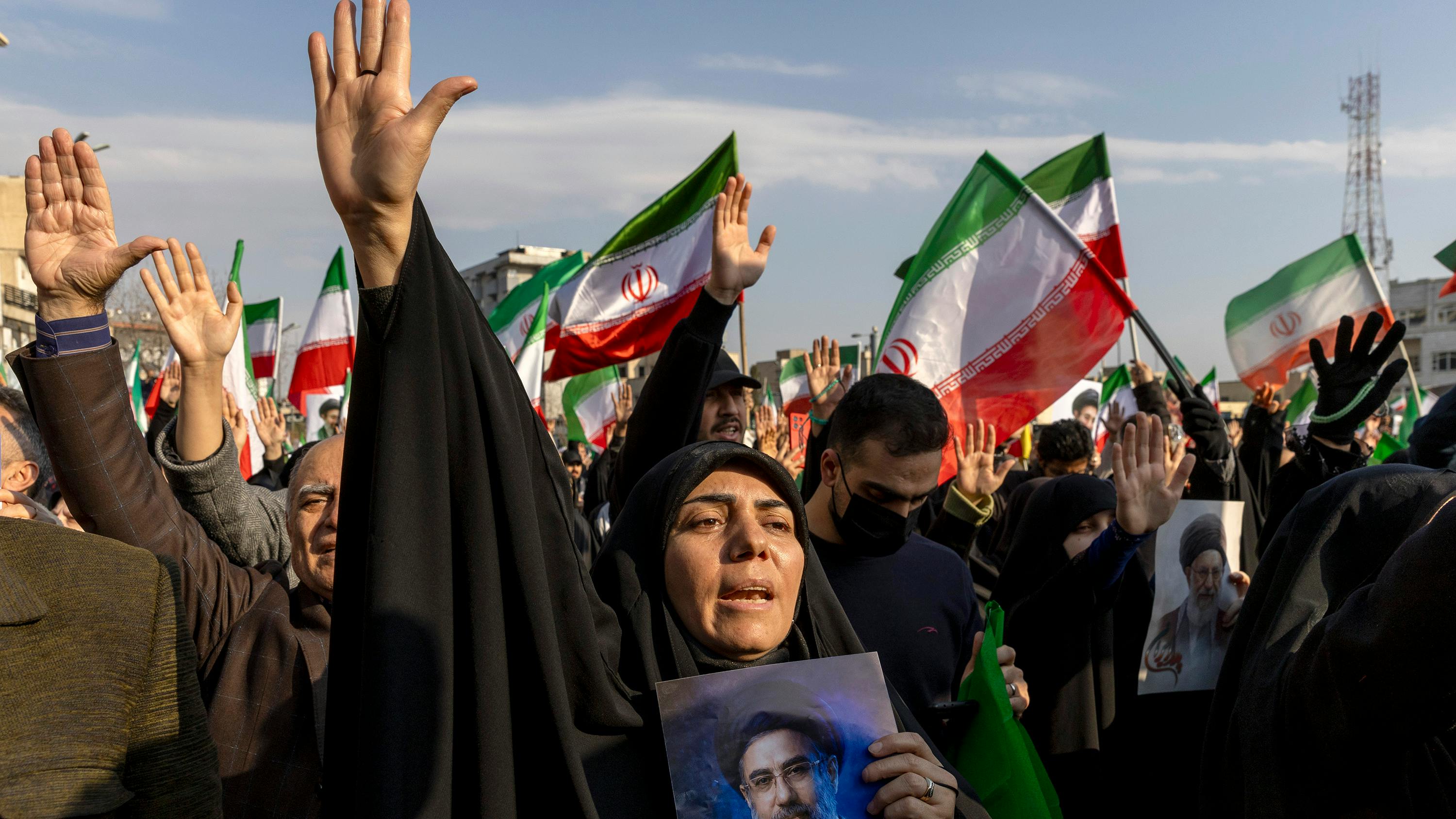 A woman holding a photograph of a man and raising her hand in the air in front of a large crowd of people raising their hands and Iranian flags