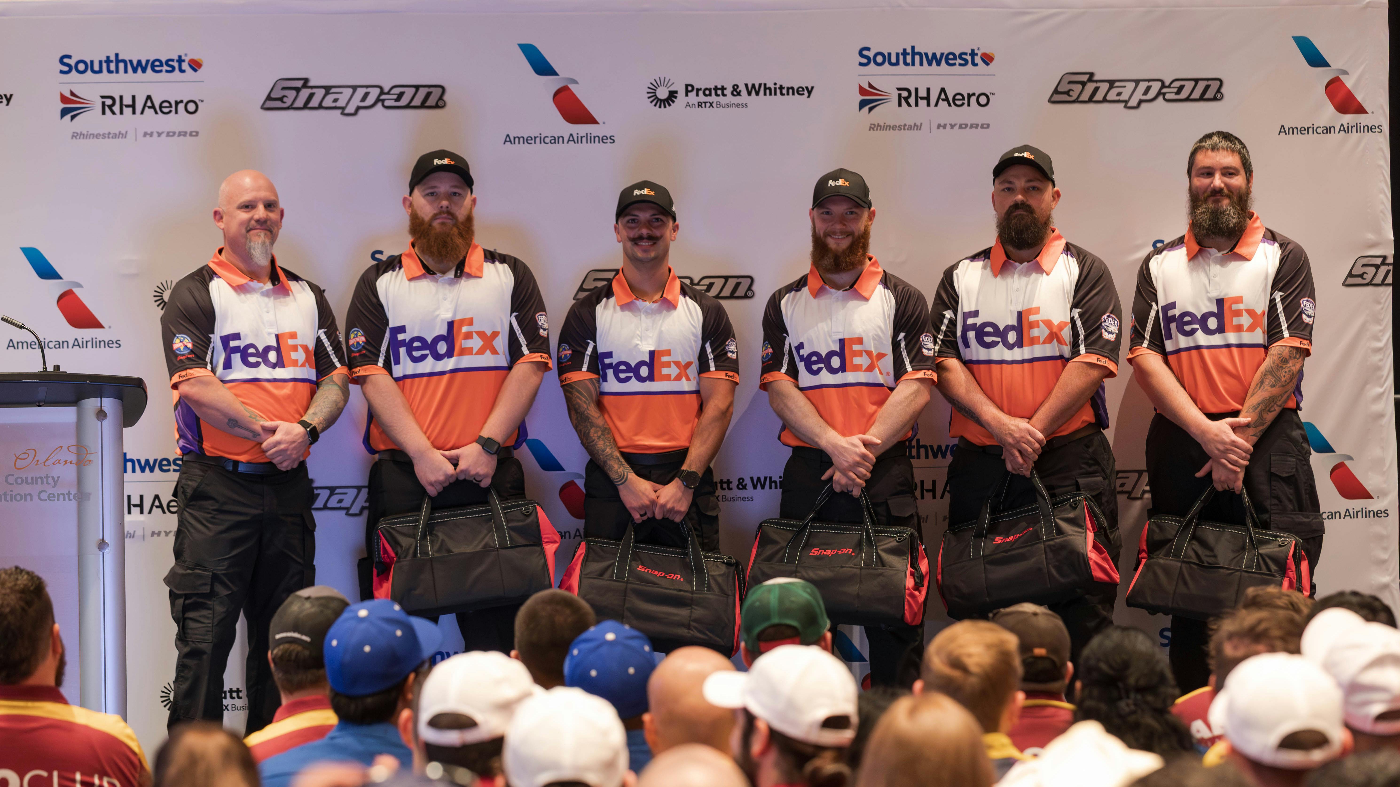 Six men wearing FedEx shirts standing in a line on a stage and smiling at the camera