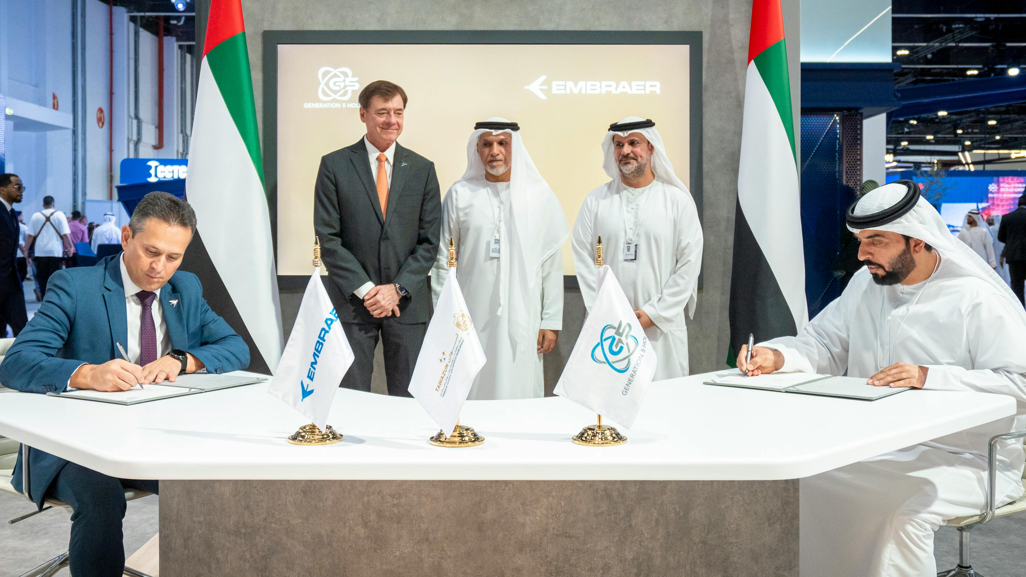 Two men sit at a table with flags on it, signing documents, while three men stand behind the table