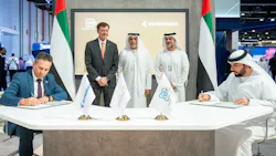 Two men sit at a table with flags on it, signing documents, while three men stand behind the table Two men sit at a table with flags on it, signing documents, while three men stand behind the table