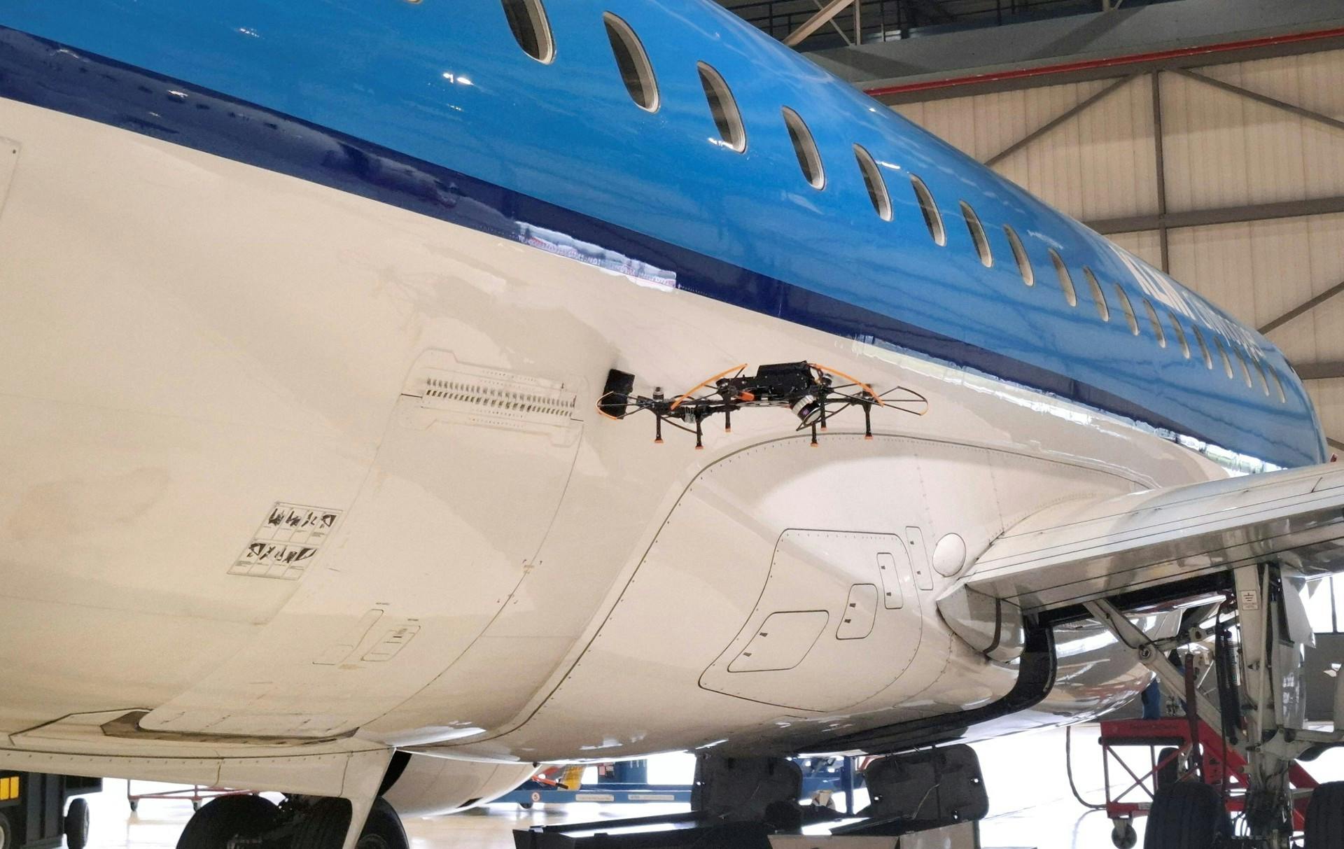 A black drone hovering next to a white and blue airplane inside a hangar